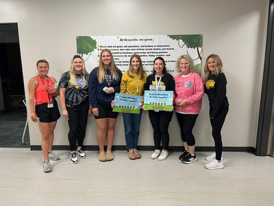 Seven teachers are standing against a wall holding two signs.