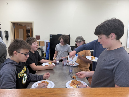 Six students eat spaghetti, with one student pouring milk in a glass.