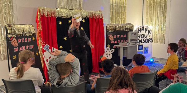 Magician performs card tricks for children sitting in a dimly lit, festive room.
