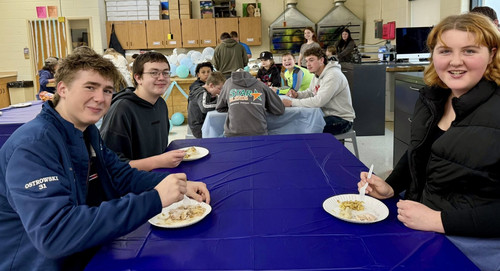High school students at a potluck event, sitting at tables and eating food from paper plates.