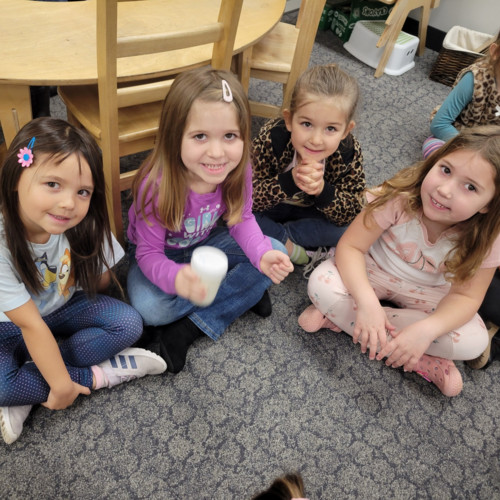 Four young elementary students sit smiling on a gray patterned carpet in a classroom.