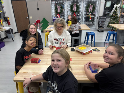 A teacher and her students smiling and playing a board game in a holiday-decorated classroom.