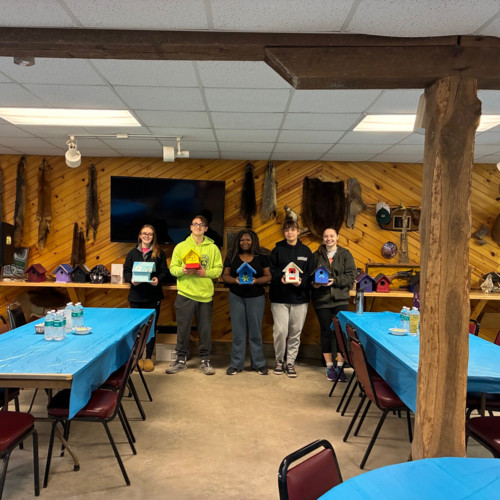 Five students displaying their hand-painted birdhouses in a wood-paneled workshop.