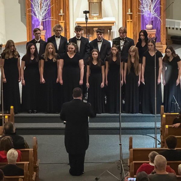 A chamber choir stands on a tiered stage in a church, performing for a seated audience.