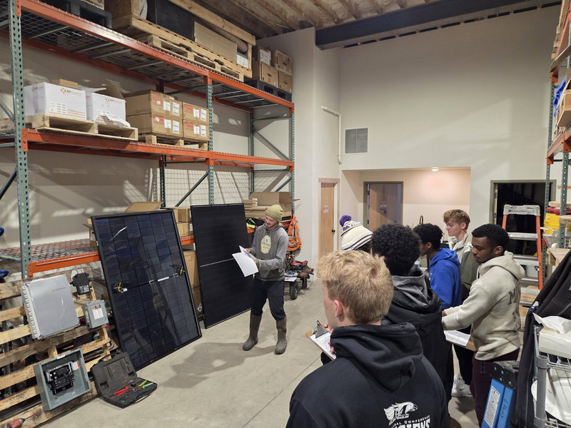 Students in a warehouse watching a demonstration of solar panels and electrical equipment.