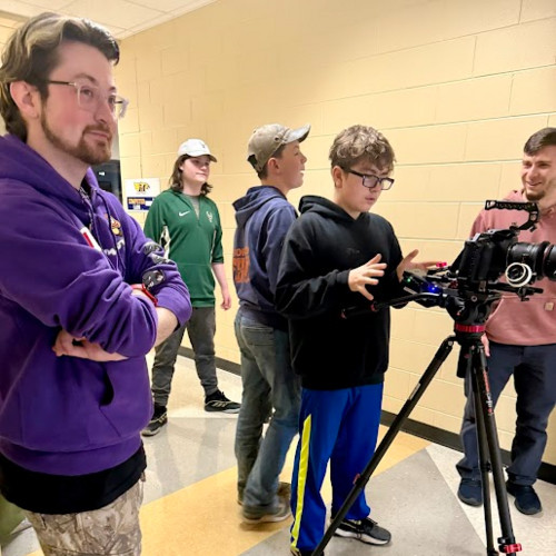 Students in a school hallway gather around a professional camera mounted on a tripod.